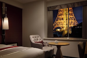 A hotel room with a bed, a chair, a table, a lamp, and a window with a view of a lit structure.