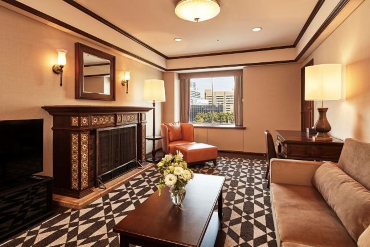 A hotel room with a fireplace, a sofa, a coffee table with flowers, and a large window with a city view.