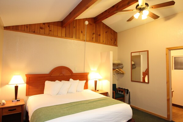 A hotel room with a wooden ceiling, a bed with white linens, two bedside lamps, a green throw blanket, a wooden nightstand, a chair, a coat rack, and a mirror on the wall. - Oakhurst Lodge (Oakhurst)