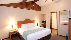 Habitación de hotel con techo de madera, cama con sábanas blancas, dos mesitas de noche con lámparas, manta verde y ventilador de techo.