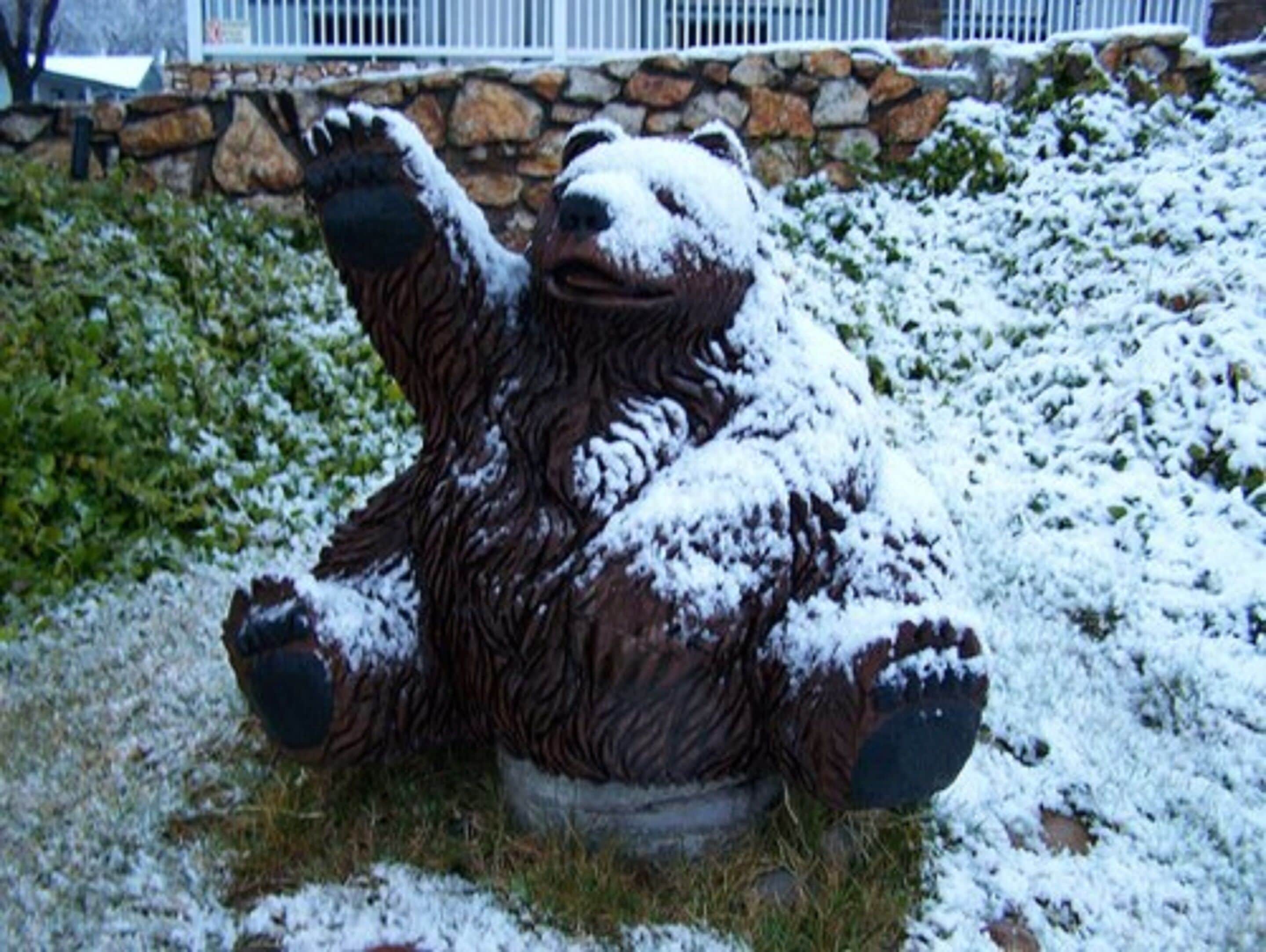 Une sculpture d’ours recouverte de neige, située dans un jardin, avec un mur de pierre et une rambarde métallique en arrière-plan.