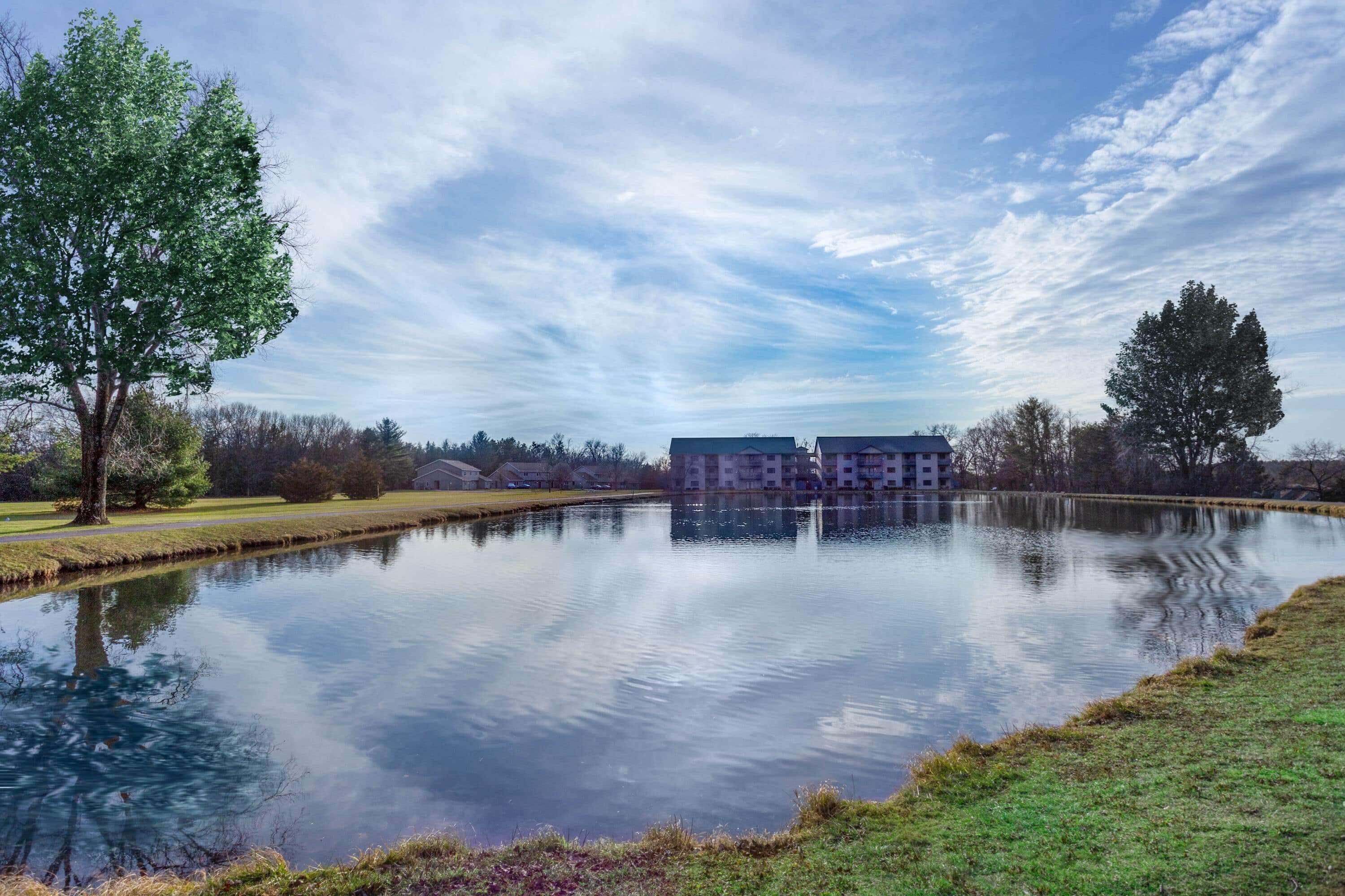 A calm lake with a reflection of trees and a building complex in the background.