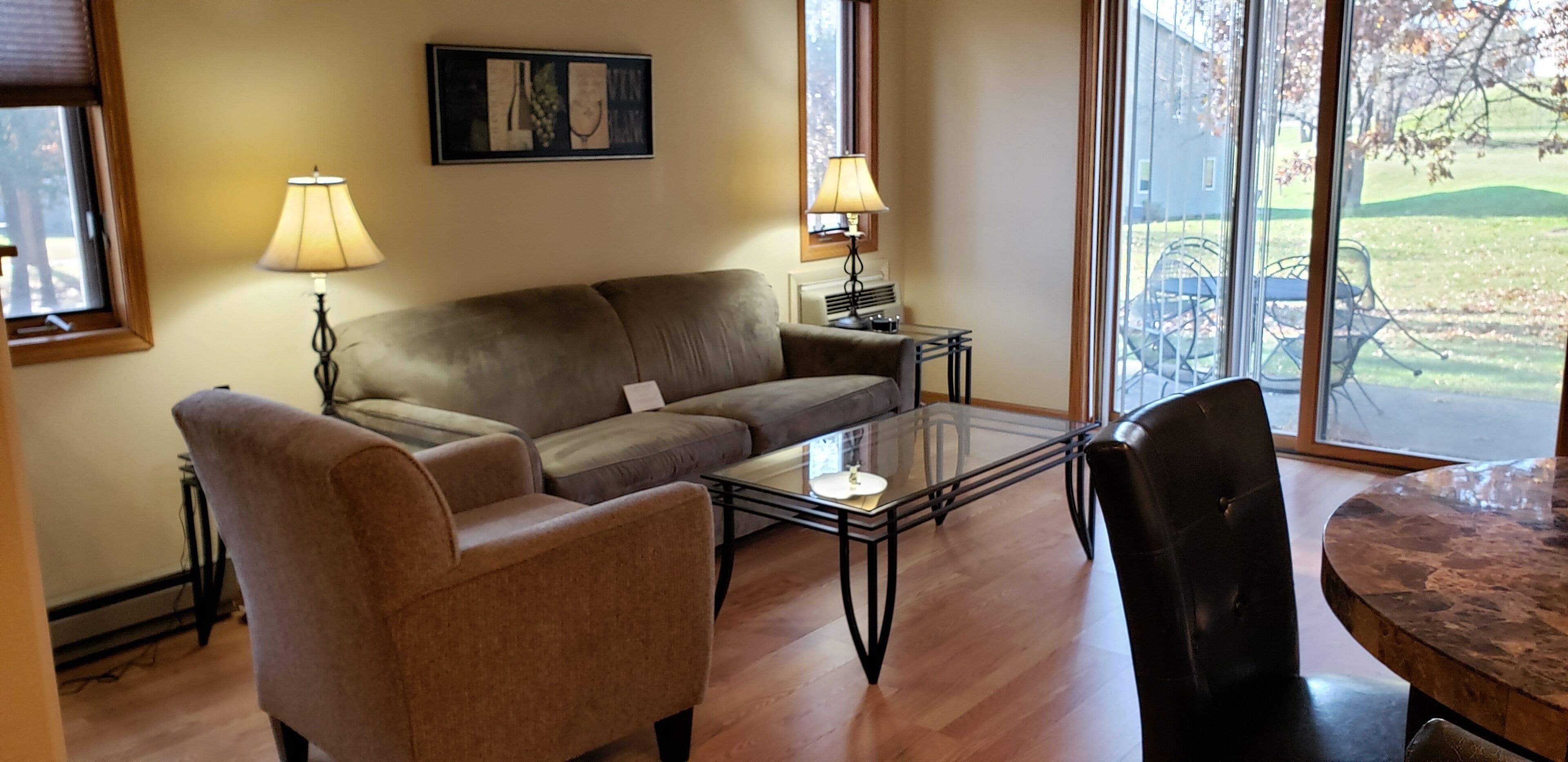 A living room with a sofa, armchair, glass coffee table, and a dining table with chairs. There are two lamps on side tables, a painting on the wall, and a view through large windows.
