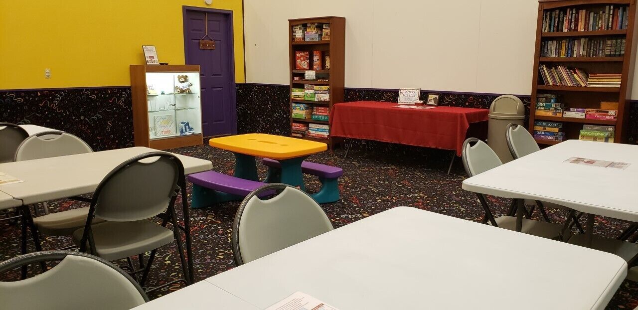 A room with tables and chairs, a bookshelf, and a table with a red tablecloth.