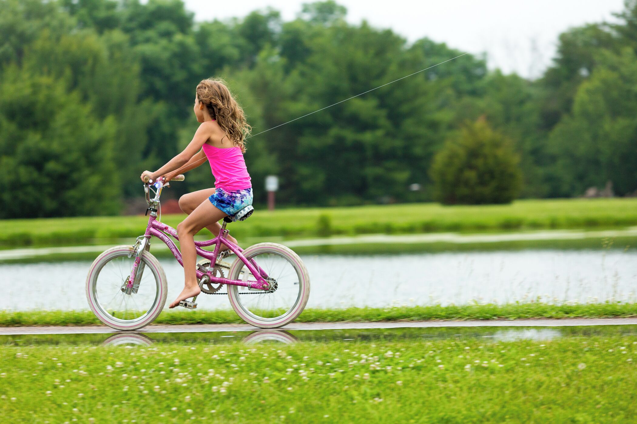 En person cyklar på en rosa cykel längs en stig intill ett vattendrag, med grön gräsmatta och träd i bakgrunden.