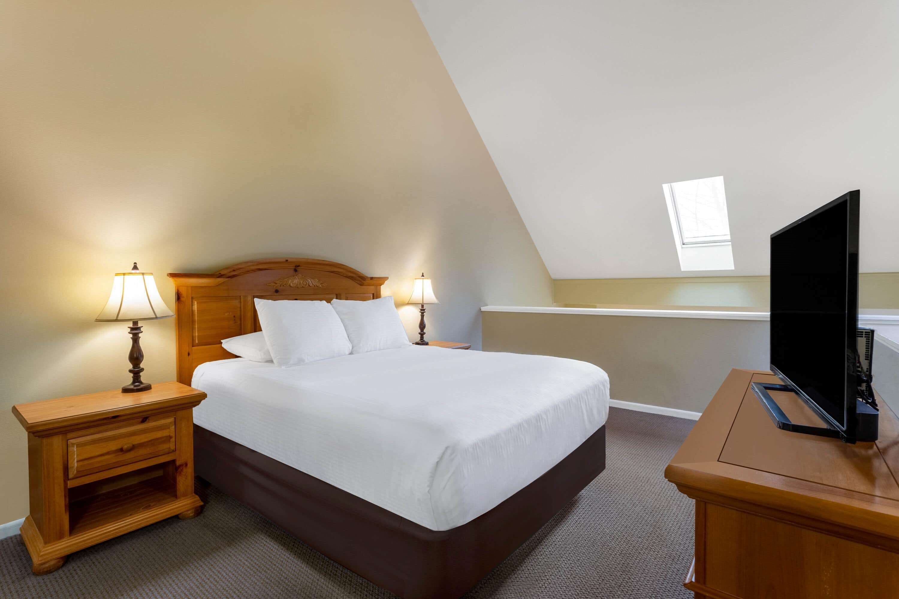 A neatly made bed in a room with a sloped ceiling, a television on a wooden stand, and two bedside lamps.
