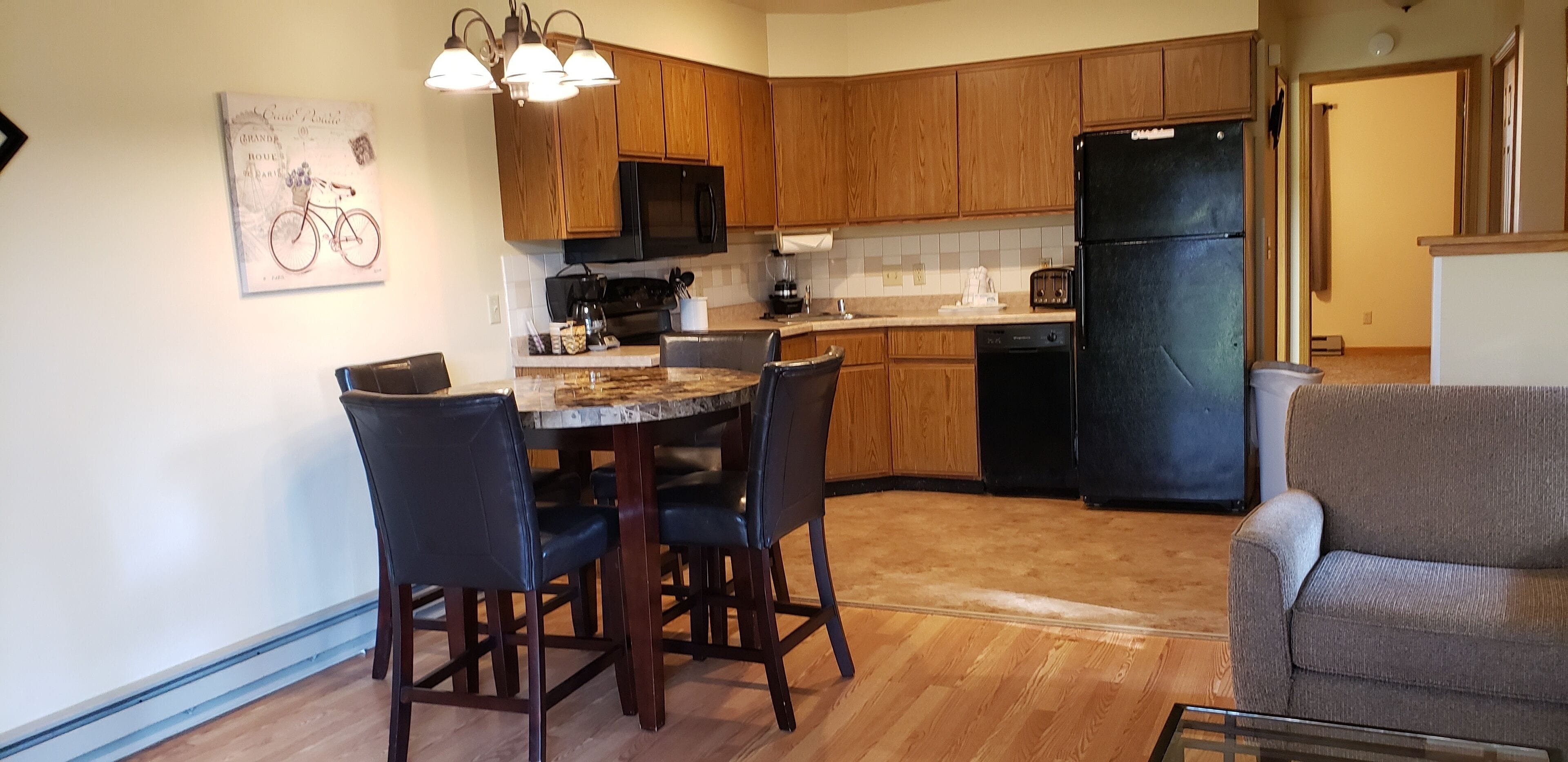 A kitchen with wooden cabinets, a black refrigerator, a dining table with chairs, and a painting on the wall.