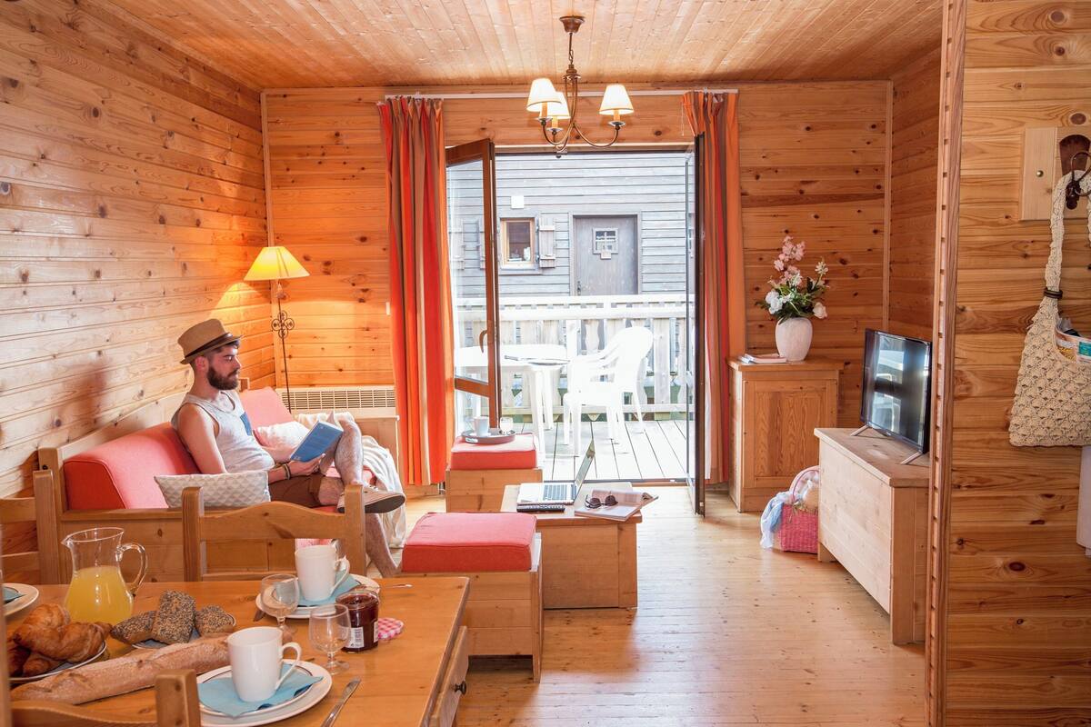 A cozy wooden cabin with a sofa, a dining table with breakfast, a TV, and a laptop.