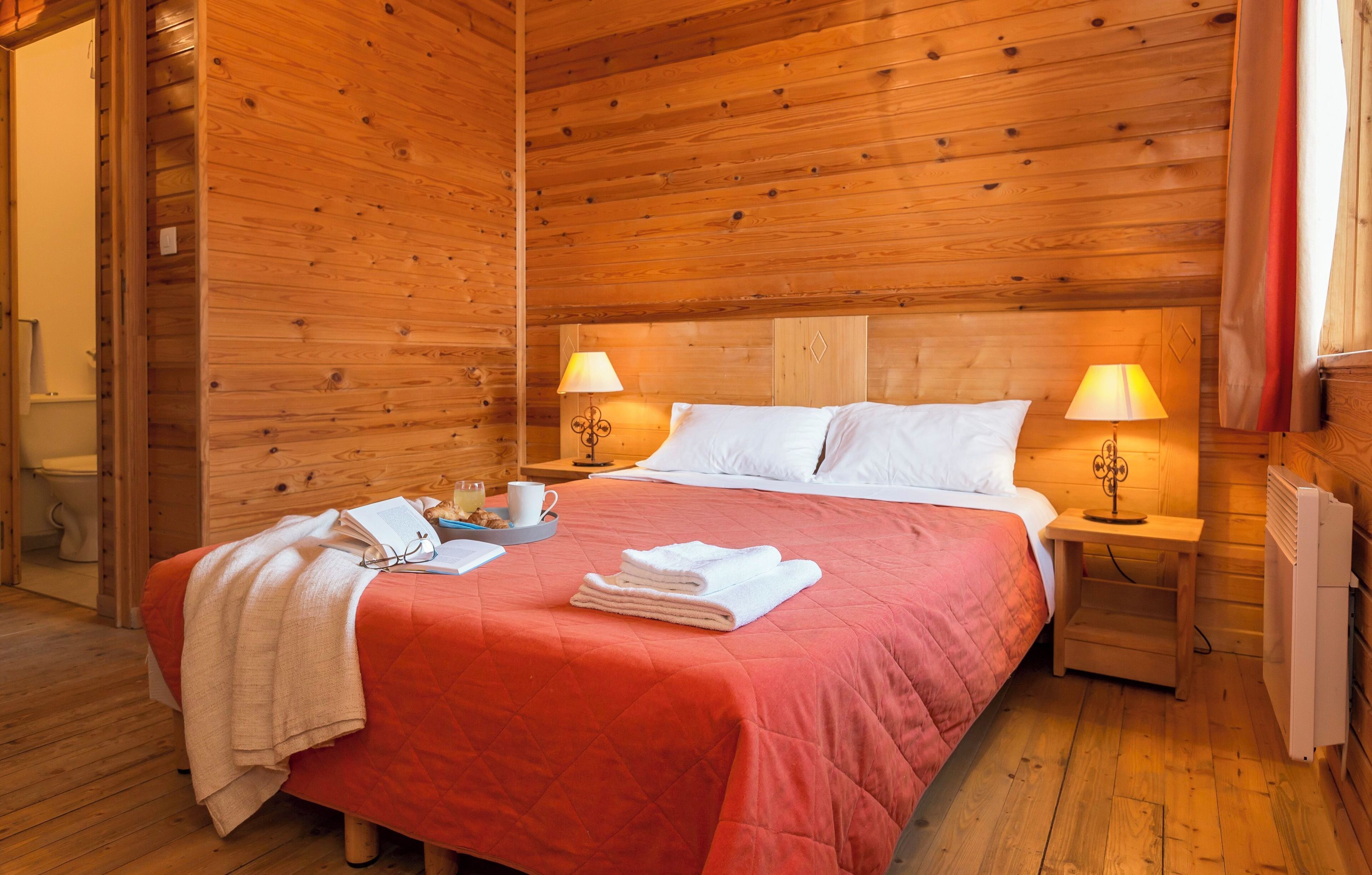 Una habitación de cabaña de madera con una cama, dos lámparas de noche, un radiador y una ventana con cortinas.