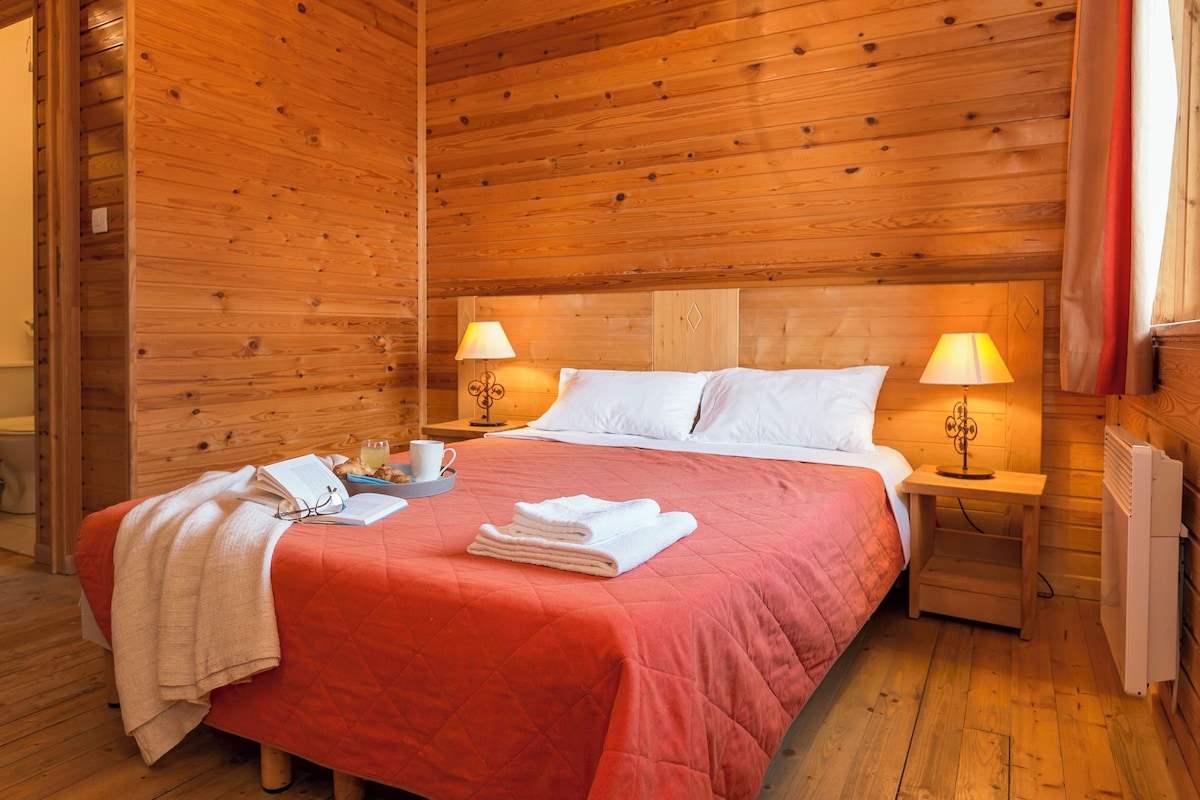 A wooden cabin room with a bed, two bedside lamps, a radiator, and a window with curtains.