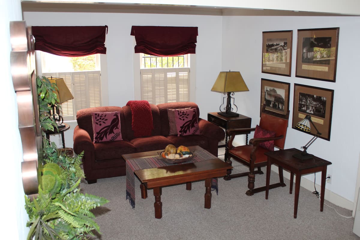 A living room with a sofa, armchair, coffee table, and a lamp. There are framed pictures on the wall and a plant in the corner.