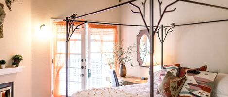 A bedroom with a four-poster bed, a fireplace, a desk with a chair, and a window with curtains.