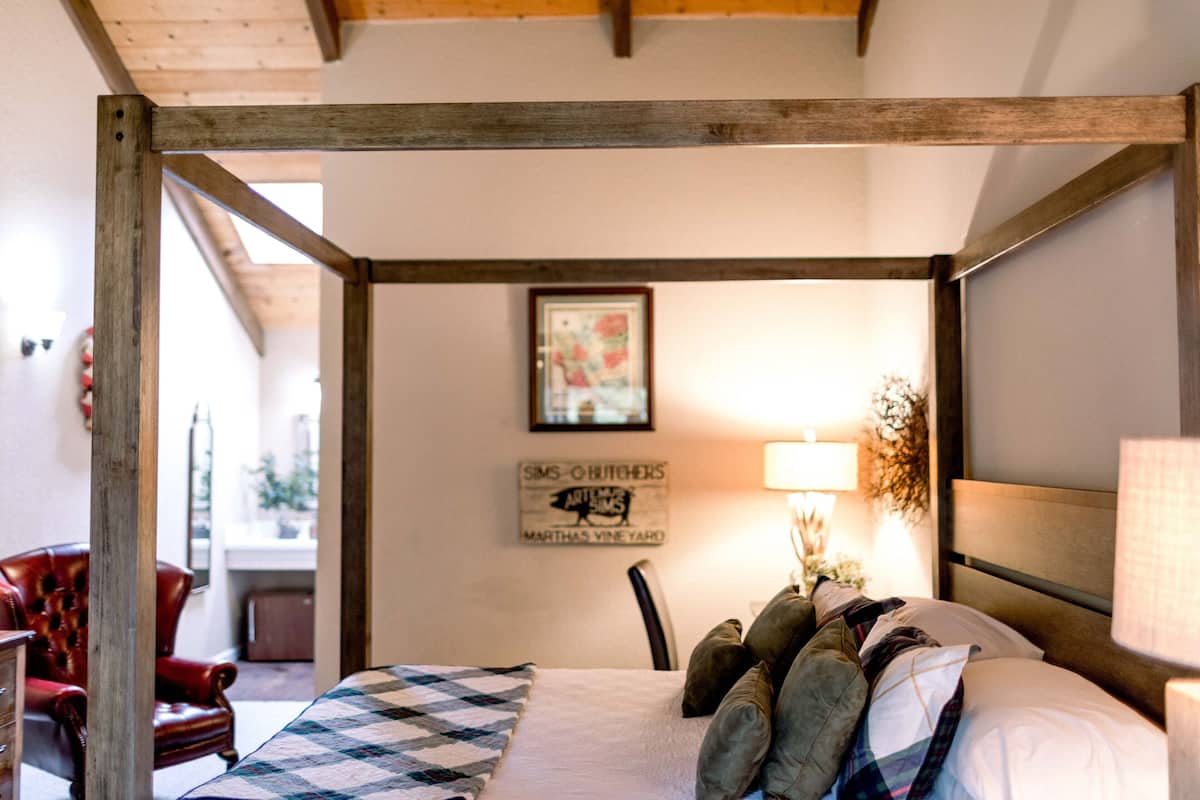 A bedroom with a wooden four-poster bed, a quilt, and a lamp. There is a chair and a sign on the wall.