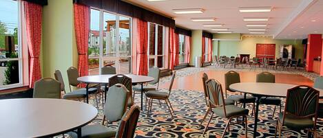 Un salón de conferencias de hotel con mesas y sillas redondas, ventanas amplias con cortinas rojas y una alfombra estampada.