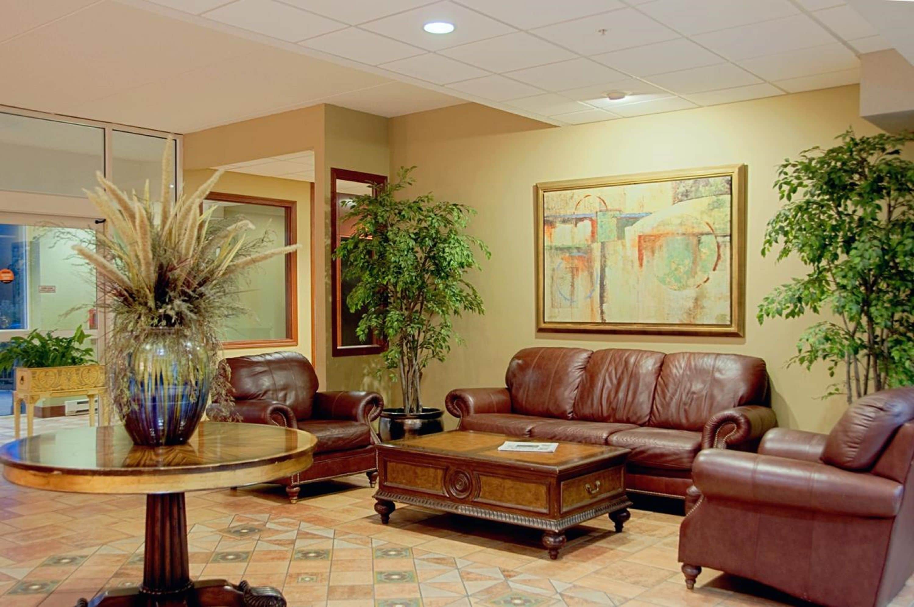 A lobby with a brown leather sofa, a round wooden coffee table, a glass-top table with a vase, and a large abstract painting on the wall.