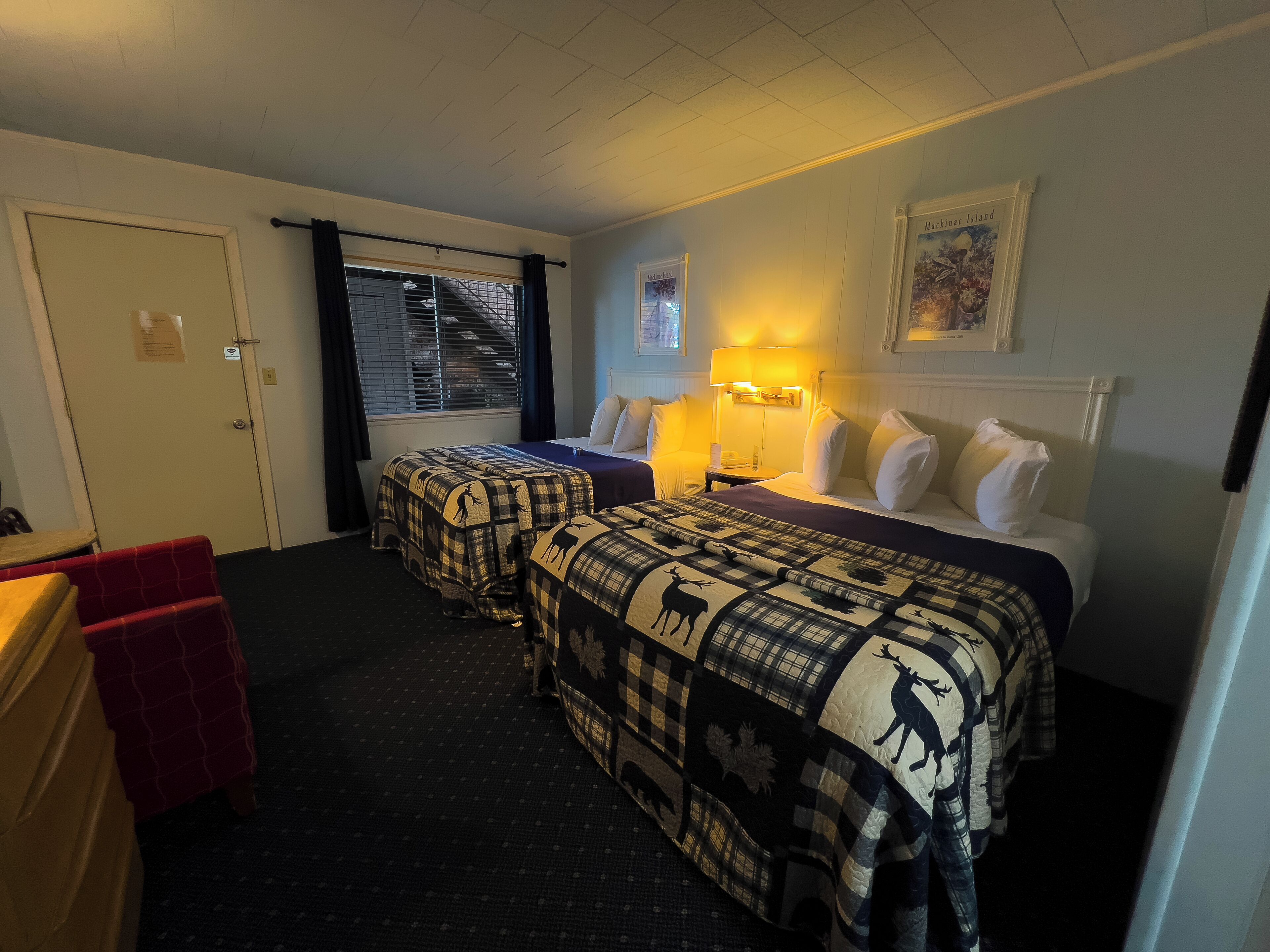 A hotel room with two beds, a desk, a chair, a window with blinds, and a framed picture on the wall.