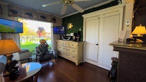 A hotel room with a wooden floor, a ceiling fan, a television, a coffee station, a white wardrobe, and a view of the outside. - American Boutique Inn - Lakeview (Mackinaw City)