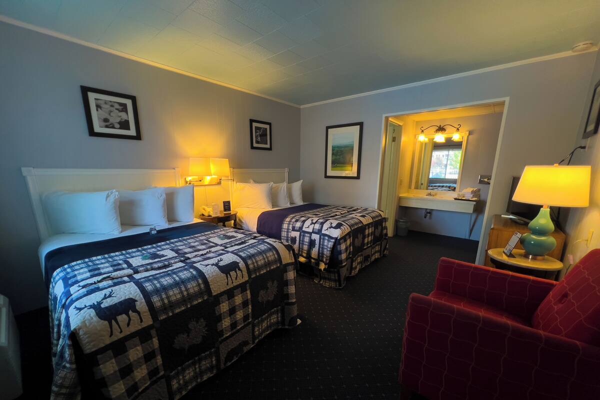Ein Hotelzimmer mit zwei Betten, einem roten Sessel, einem kleinen Tisch mit einer Lampe und einem durch eine offene Tür einsehbaren Badezimmer.