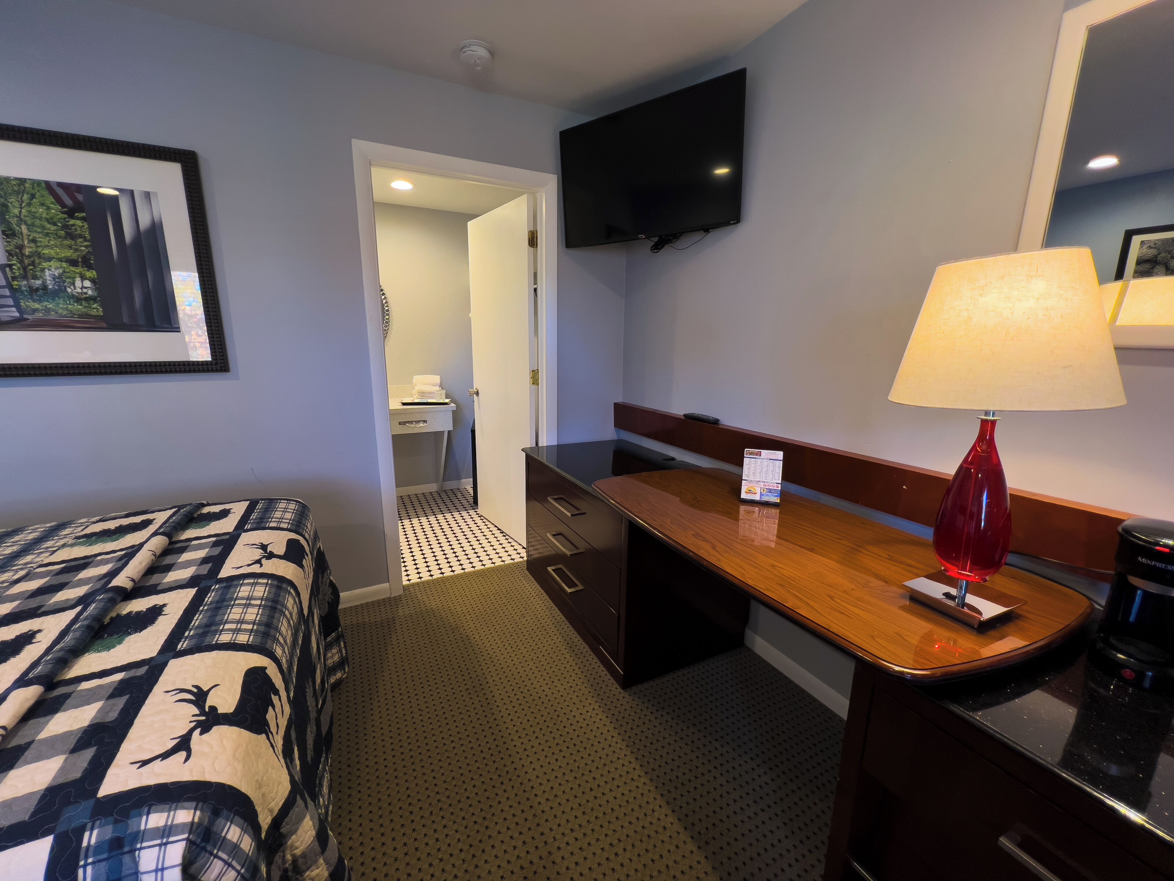 A hotel room with a bed, a desk, a lamp, a television, and a framed picture on the wall.