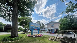 An American Boutique Inn sign with a lawn and flowers, a large tree, and a building with a brick facade. - American Boutique Inn - Lakeview (Mackinaw City)