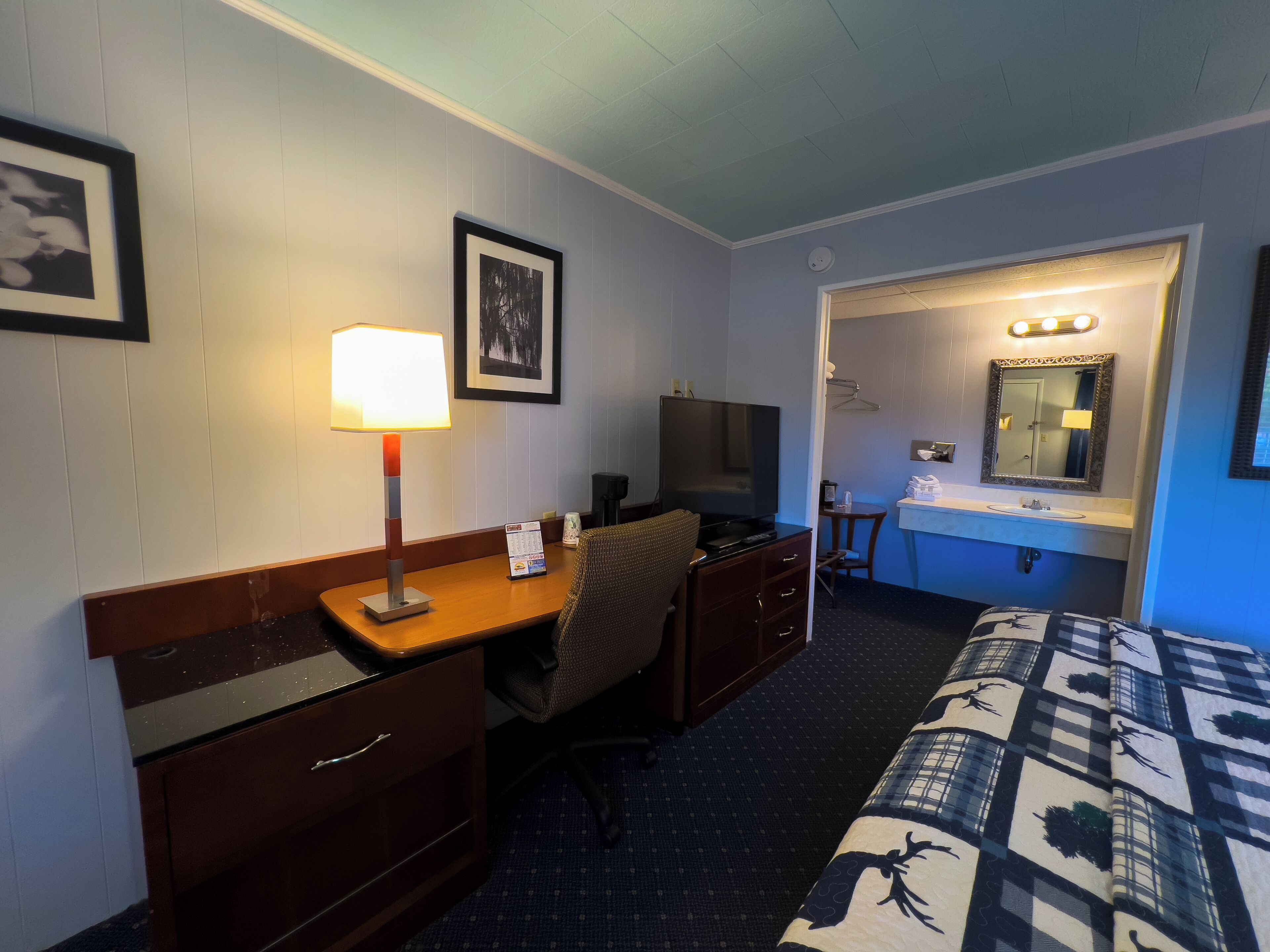 A hotel room with a bed, a desk with a lamp, a TV, a dresser, and a bathroom with a mirror and sink.