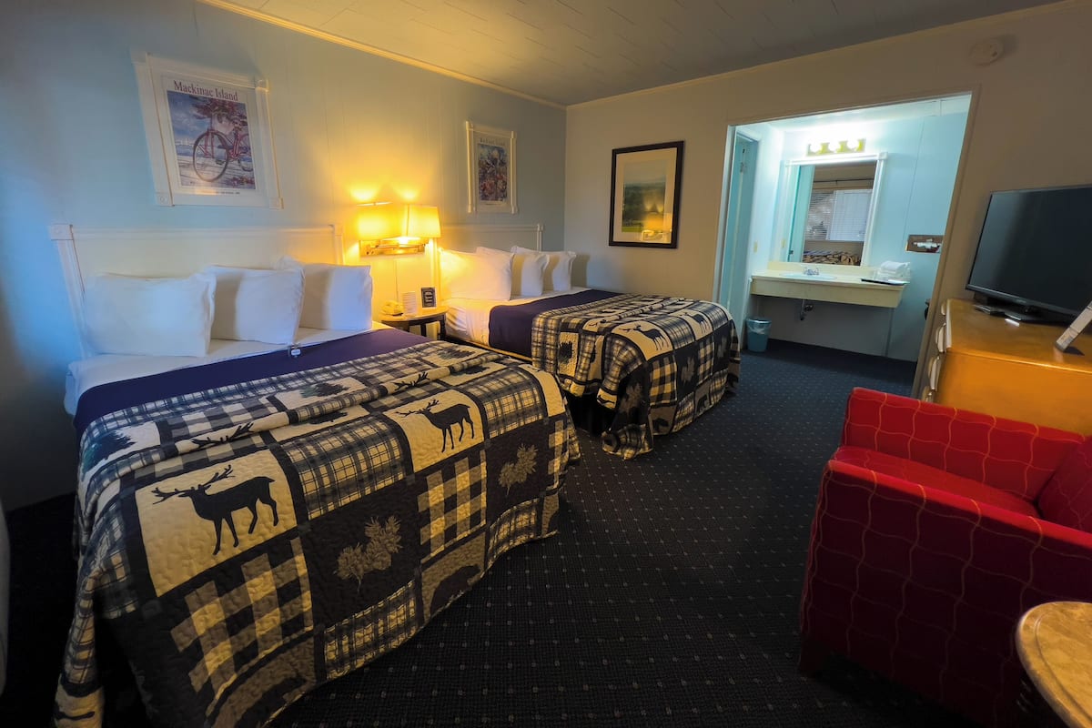 Ein Hotelzimmer mit zwei Betten, einem roten Sessel, einem Fernseher, einem Schreibtisch mit Spiegel und gerahmten Bildern an der Wand.