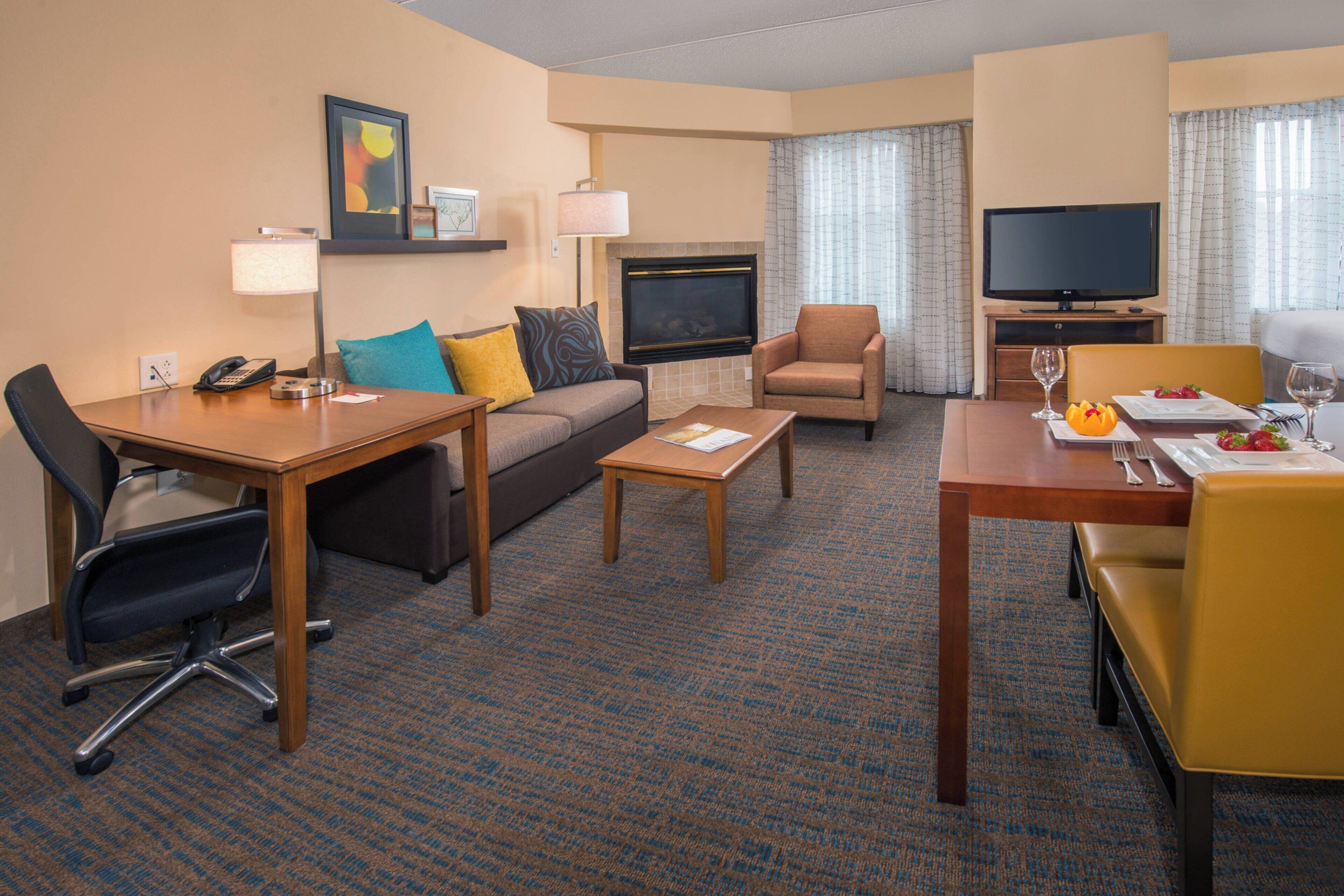 A hotel room with a sofa, armchair, dining table, television, fireplace, and a carpeted floor.
