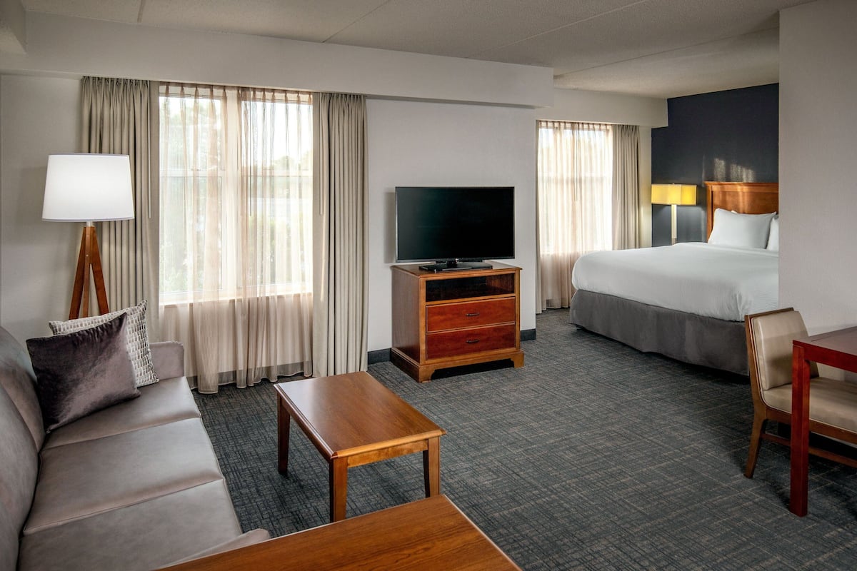 A hotel room with a bed, a sofa, a television on a wooden stand, a desk, and a lamp.