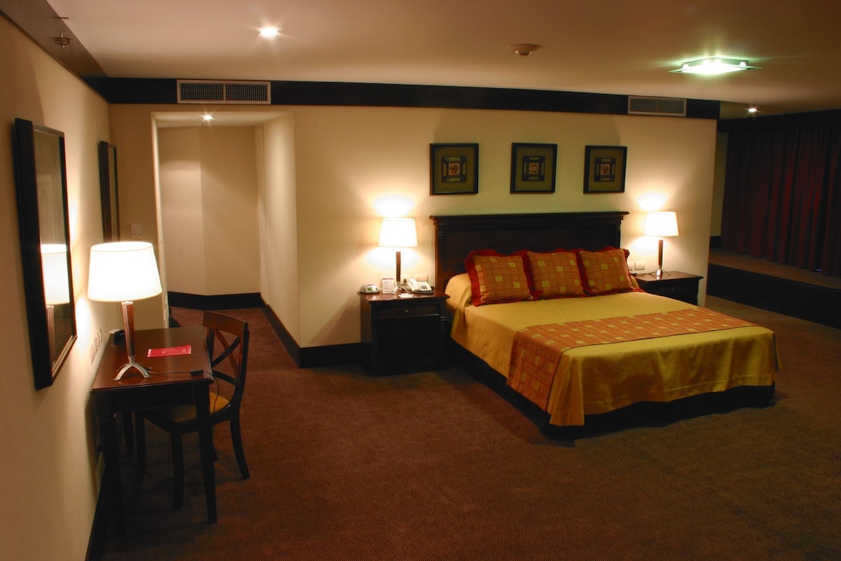 Ein Hotelzimmer mit einem Bett, zwei Nachttischen mit Lampen, einem Schreibtisch mit Stuhl und einer Tür zu einem anderen Raum.