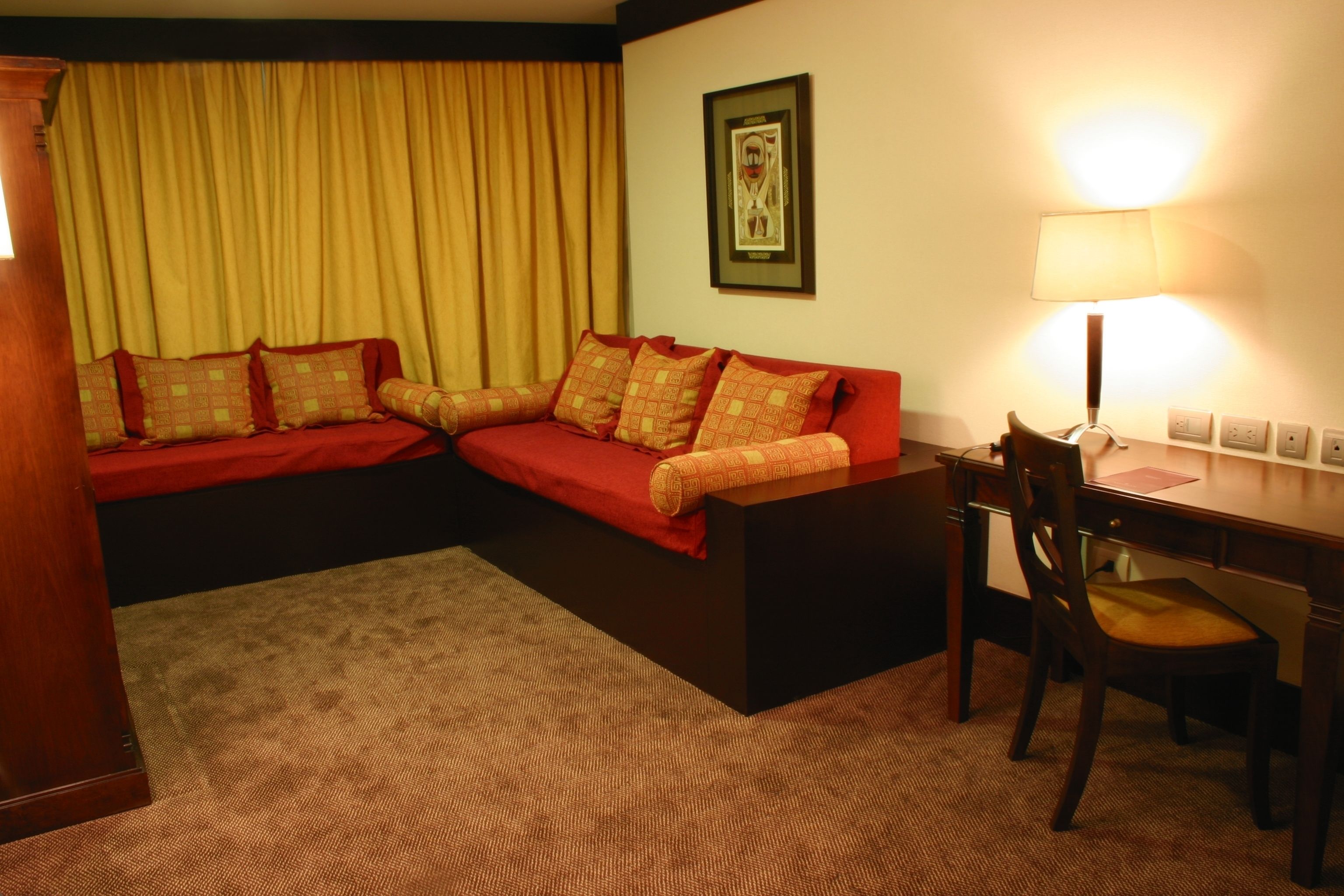 A hotel room with a red sofa set, a wooden desk with a chair, a lamp, and a framed picture on the wall.