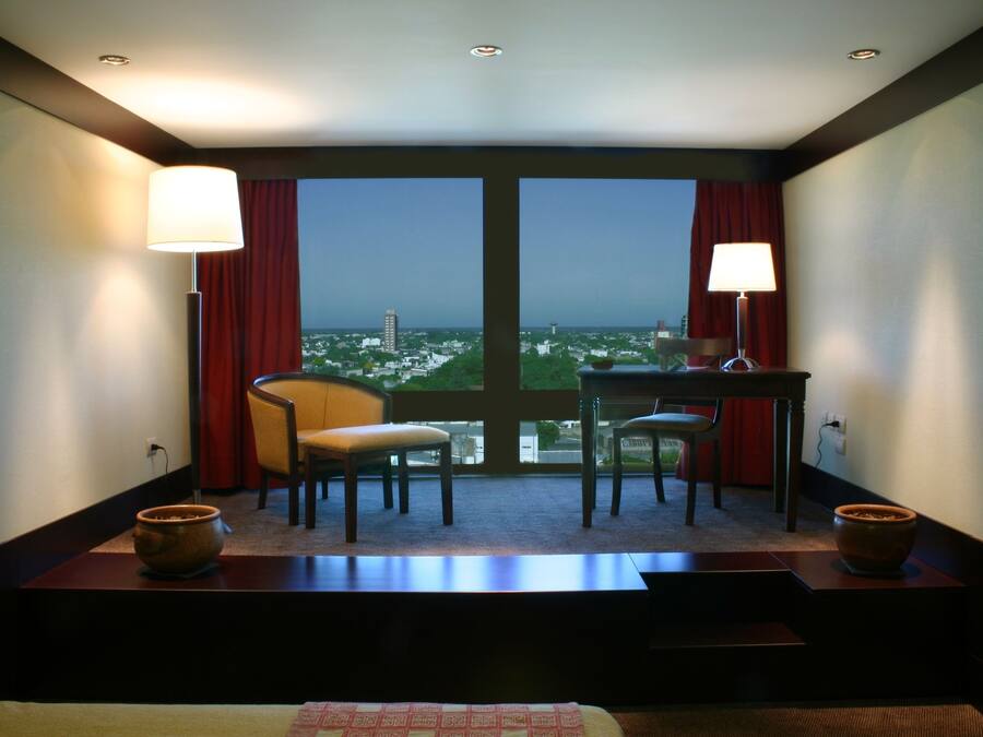 A hotel room with a large window, two chairs, a small table, and two lamps.