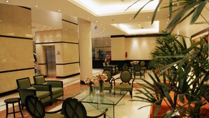 A lobby with a glass coffee table surrounded by green upholstered chairs, a reception desk, and a large potted plant. - Amérian Hotel Casino Gala (Resistencia)