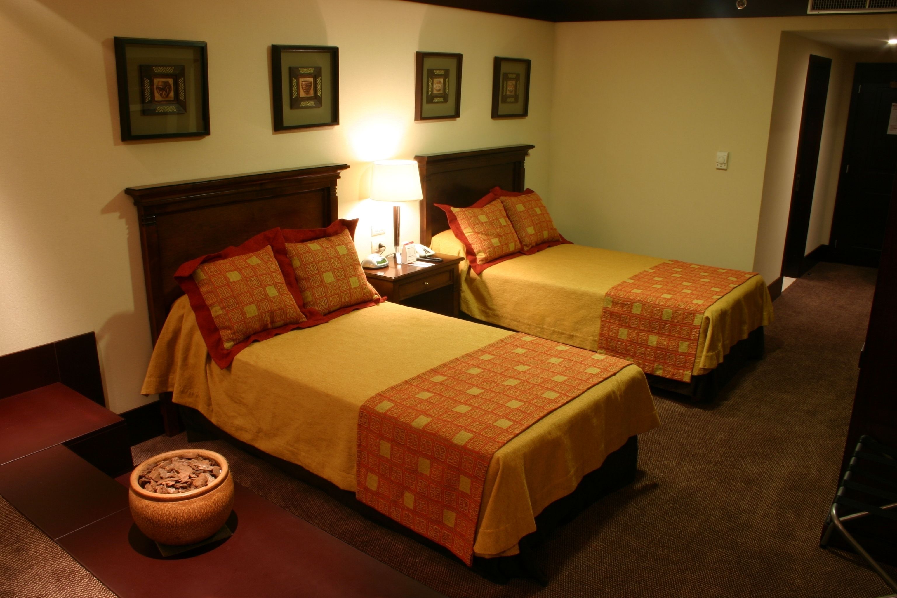 A hotel room with two beds, a nightstand with a lamp, a small table with a bowl, and framed pictures on the wall.