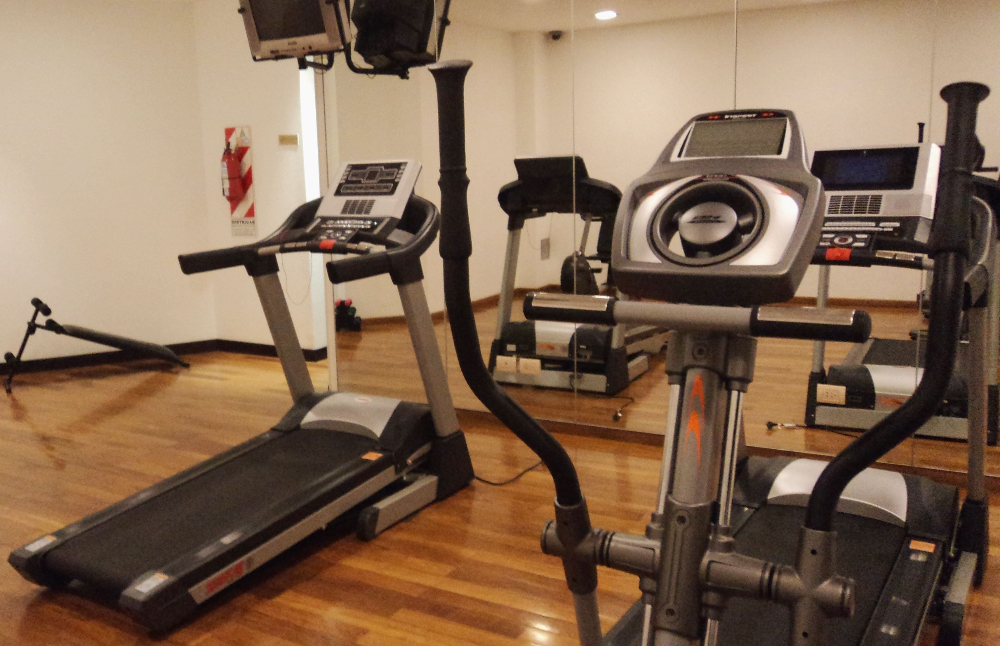 A gym with various exercise machines, including treadmills and elliptical trainers, on a wooden floor.