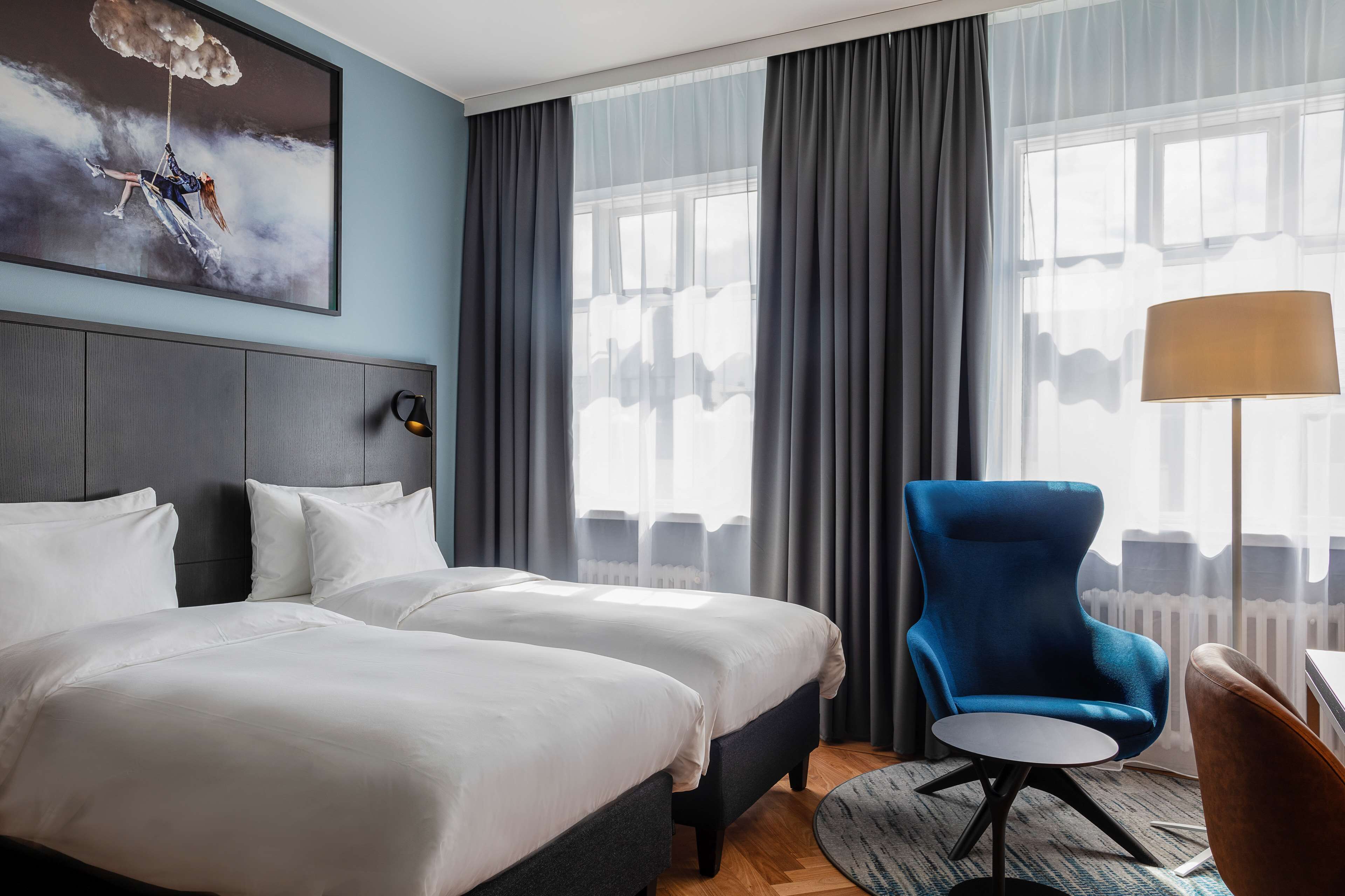 Ett hotellrum med två sängar, en blå fåtölj, ett litet bord och ett stort fönster med gardiner.