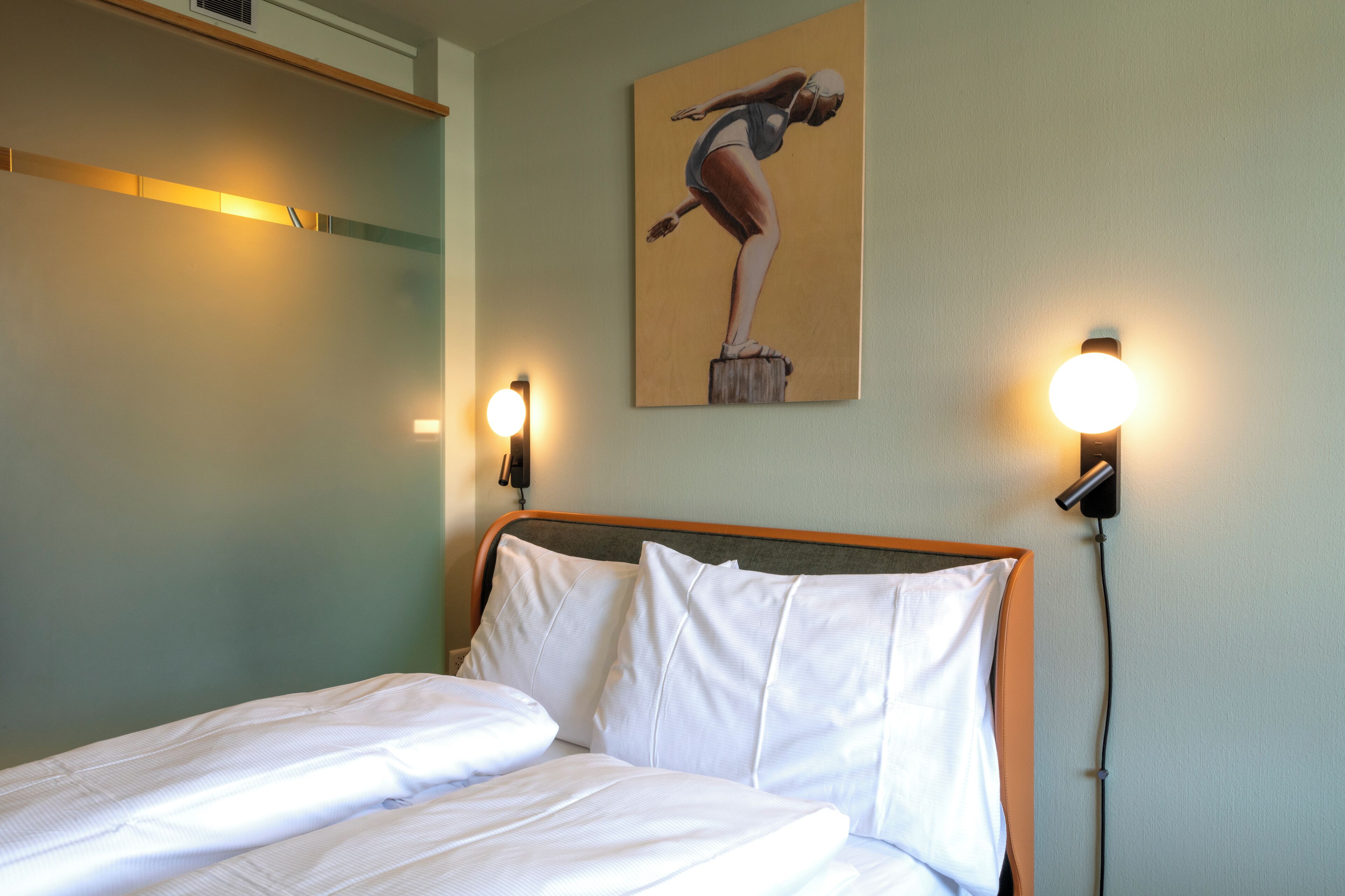 Una cama bien hecha con sábanas blancas, un cabecero de madera y dos lámparas de lectura fijadas a la pared.