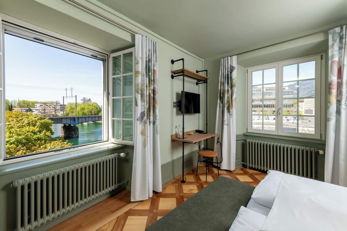 A room with a bed, a desk with a chair, a TV, and large windows with curtains.