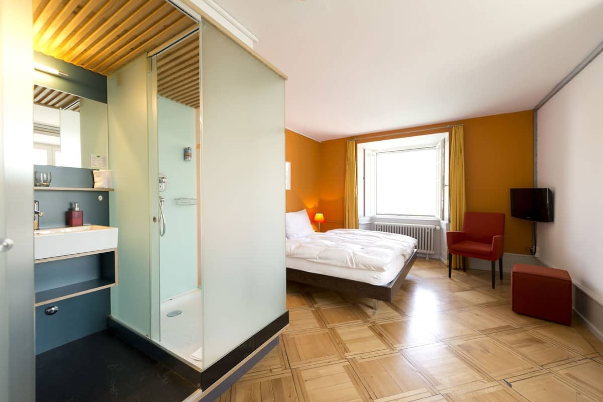 A hotel room with a bed, a desk with a chair, a TV, a window with curtains, and a bathroom with a shower and sink.