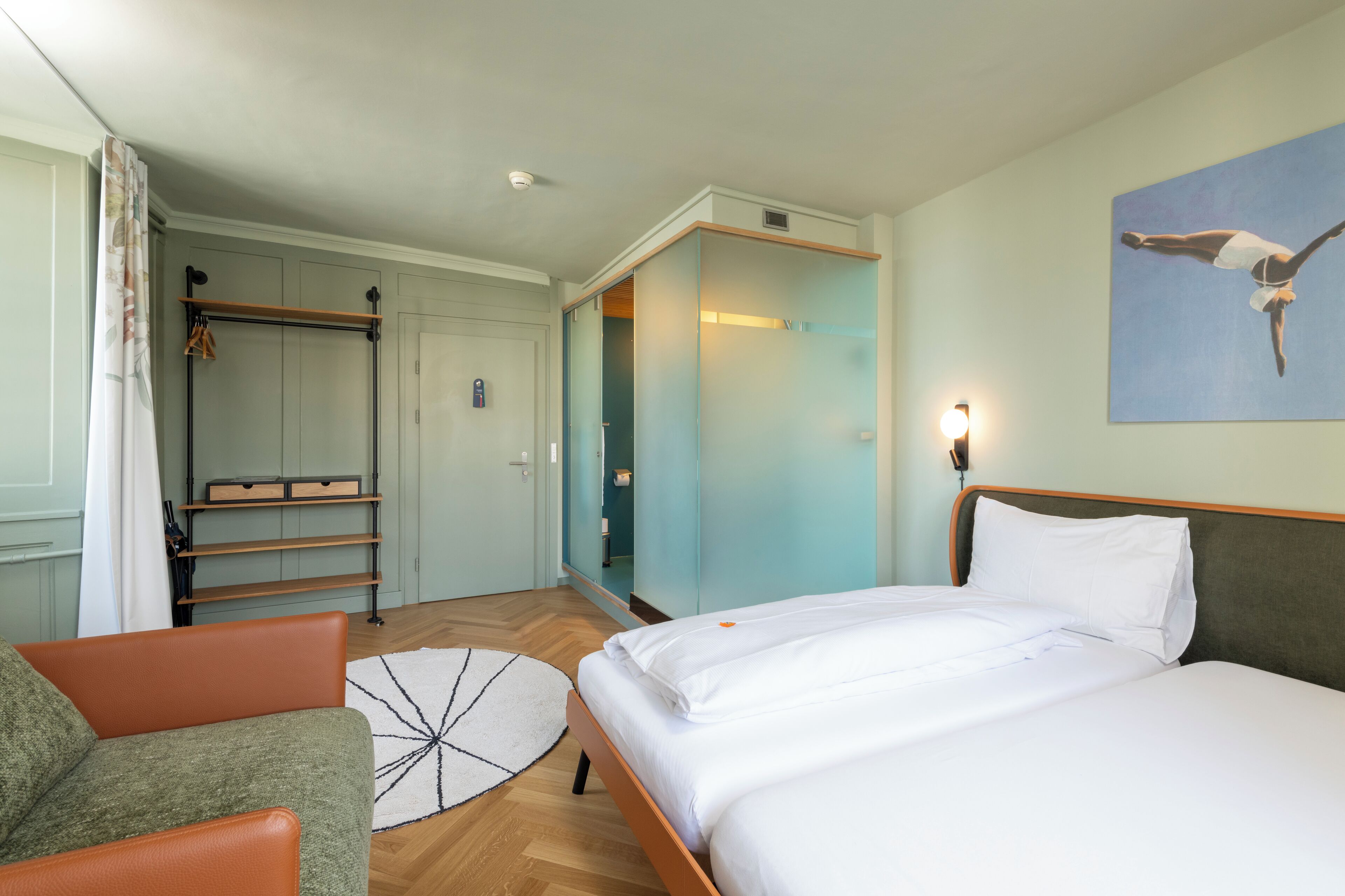 A hotel room with a bed, a chair, a wardrobe, and a painting on the wall.