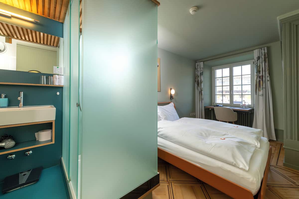 A hotel room with a bed, a desk, a chair, a bathroom with a mirror, and a window with curtains.