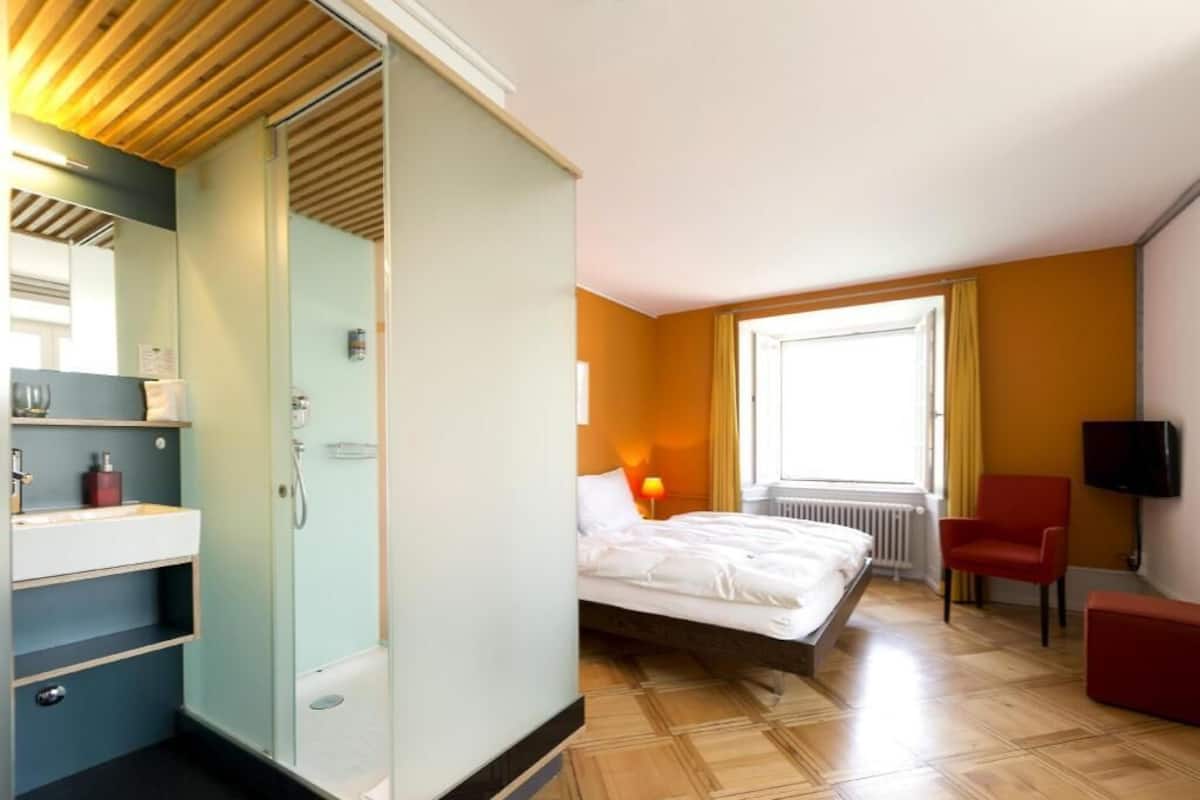 A hotel room with a bed, a shower, a TV, a red chair, and a window with curtains.