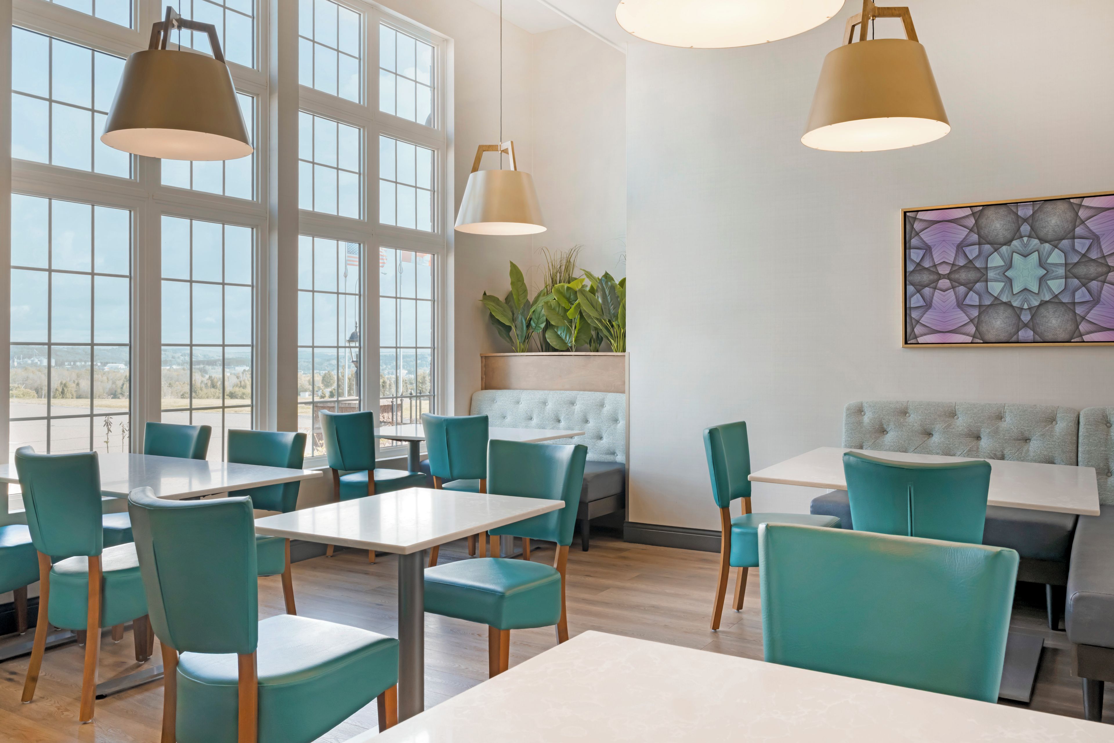 Un interior de restaurante con ventanas amplias, sillas color turquesa, mesas blancas y un banco capitonado.
