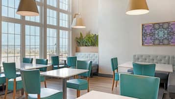 Un interior de restaurante con ventanas amplias, sillas color turquesa, mesas blancas y un banco capitonado.