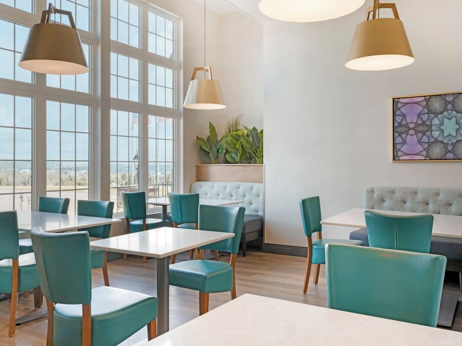 Un interior de restaurante con ventanas amplias, sillas color turquesa, mesas blancas y un banco capitonado.