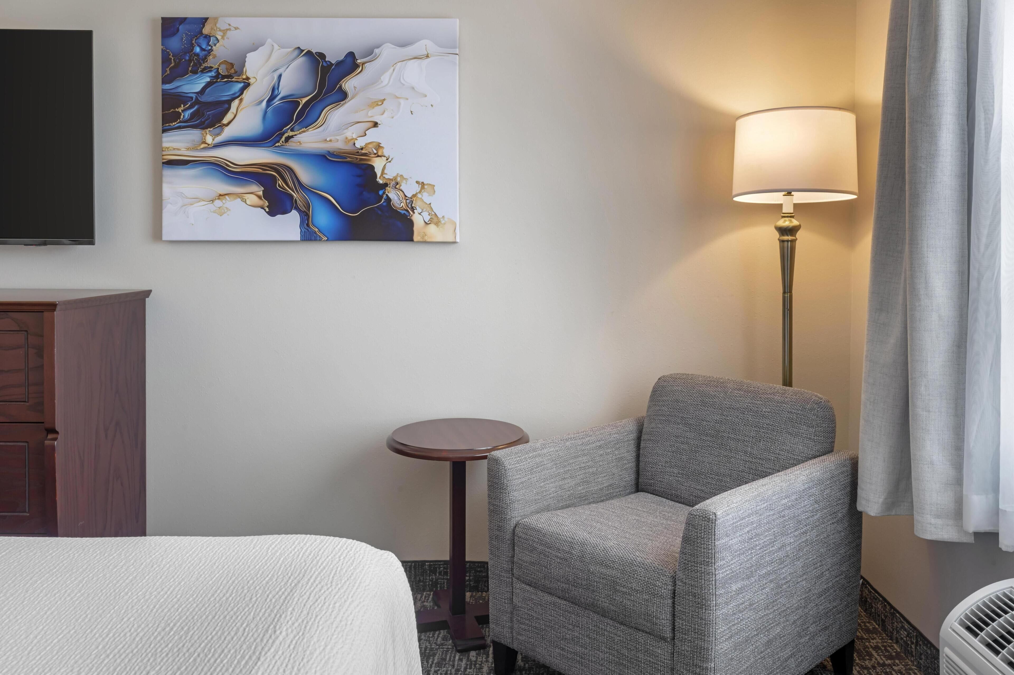 Ein Hotelzimmer mit einem Bett, einem grauen Sessel, einem kleinen runden Tisch, einer Lampe und einem Bild an der Wand.