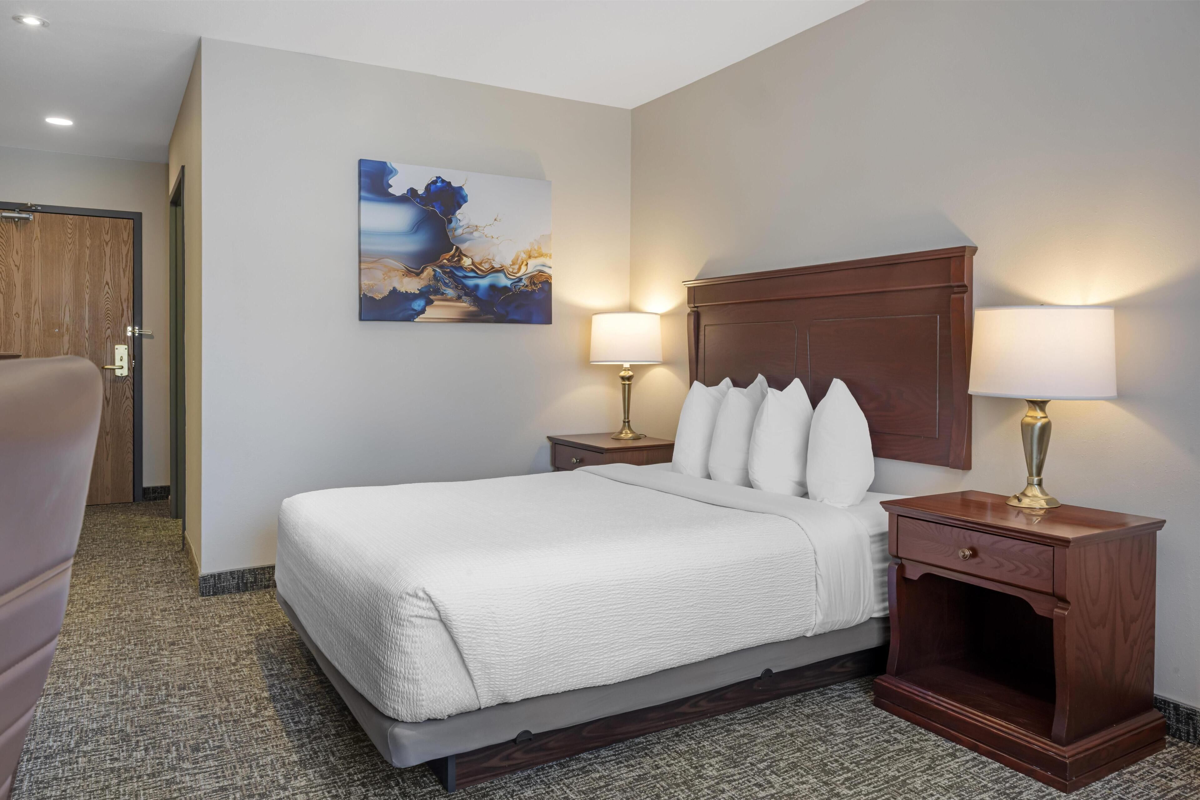 A hotel room with a large bed, two bedside tables with lamps, a wooden headboard, a painting on the wall, and a carpeted floor.