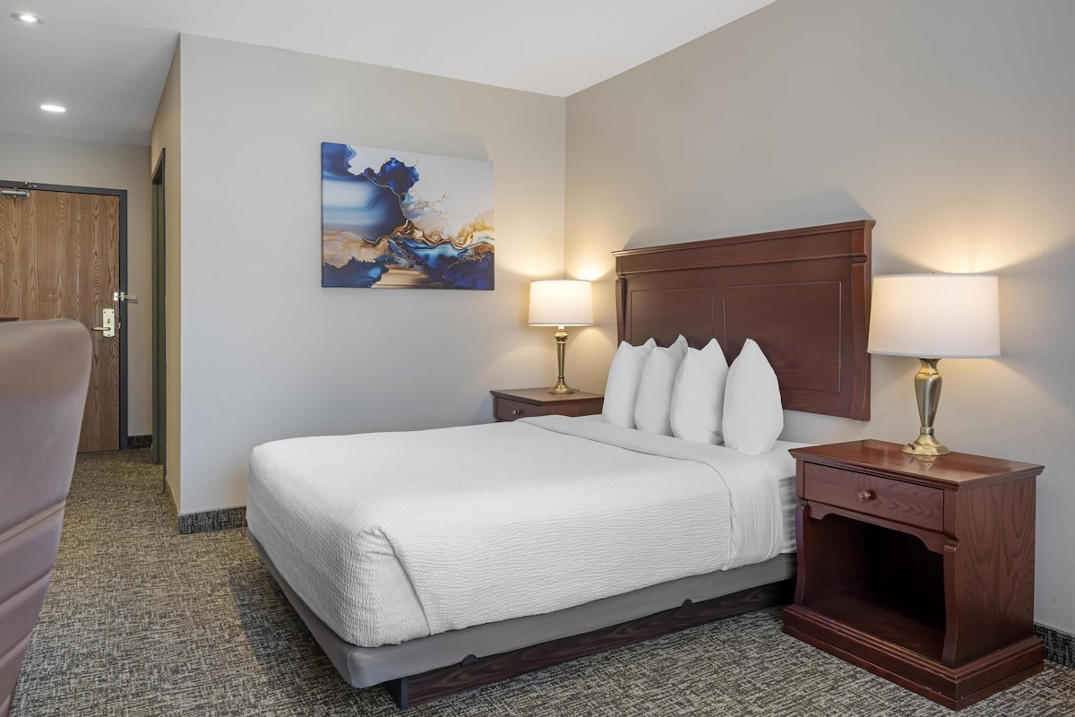 Una camera d'albergo con un letto grande, due comodini con lampade, una testiera in legno, un quadro appeso al muro e un pavimento rivestito di moquette.