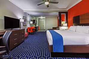 Ein Hotelzimmer mit einem großen Bett, einem Schreibtisch, einem Flachbildfernseher, einem Sessel, einer Schreibtischlampe und einem Deckenventilator.