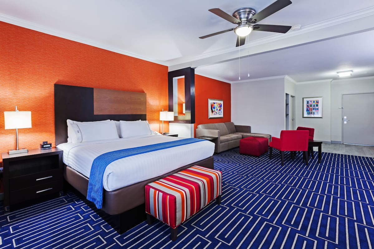 A hotel room with a large bed, a ceiling fan, a red armchair, a red ottoman, and a blue patterned carpet.