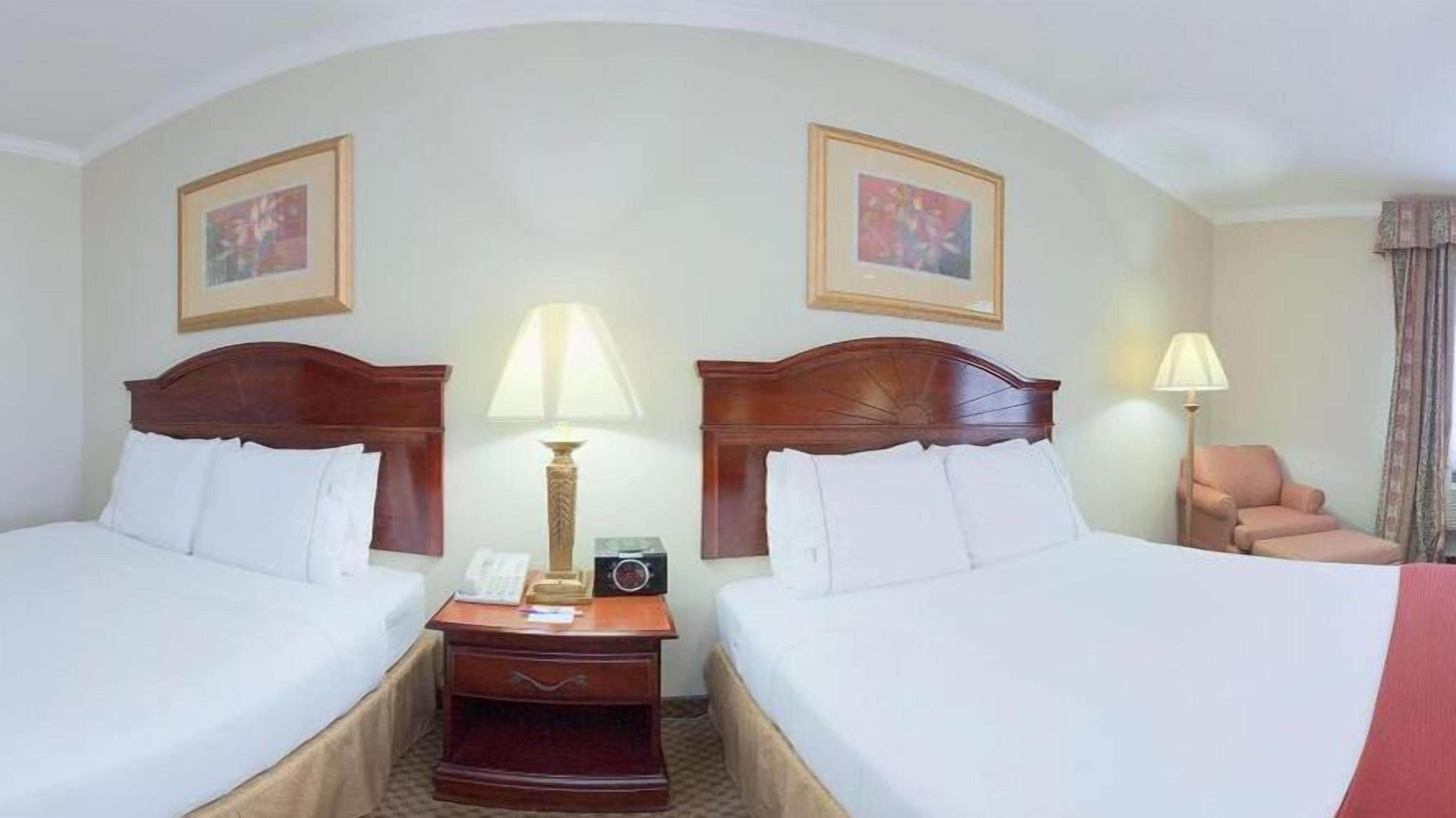 Une chambre d’hôtel avec deux lits, une table de chevet avec une lampe, une chaise et deux tableaux encadrés au mur.